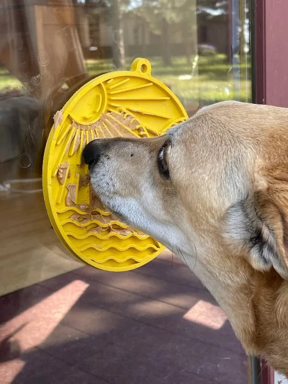Dog licking a yellow Sun n' Sea enrichment lick mat with a beach design, secured on a glass surface.