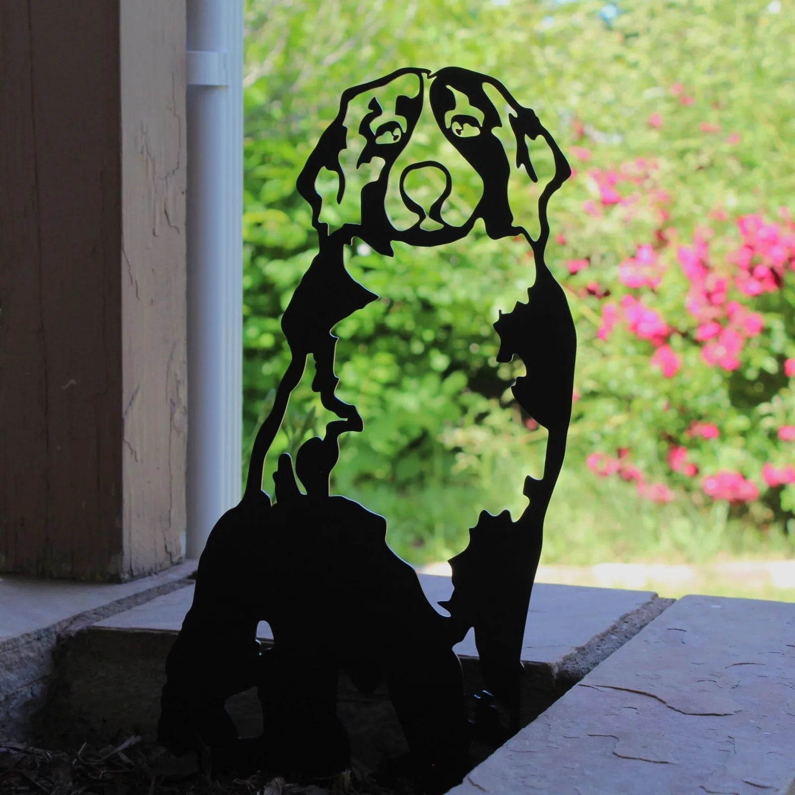 Black silhouette of a Swiss Mountain Dog made from sturdy metal, standing against a vibrant green and pink garden backdrop.