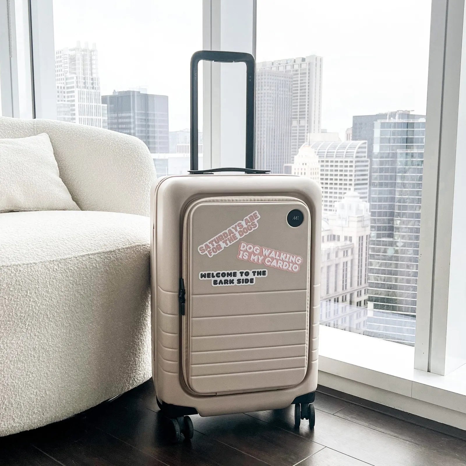 A light beige suitcase featuring playful dog-themed stickers on a windowsill with a cityscape backdrop.