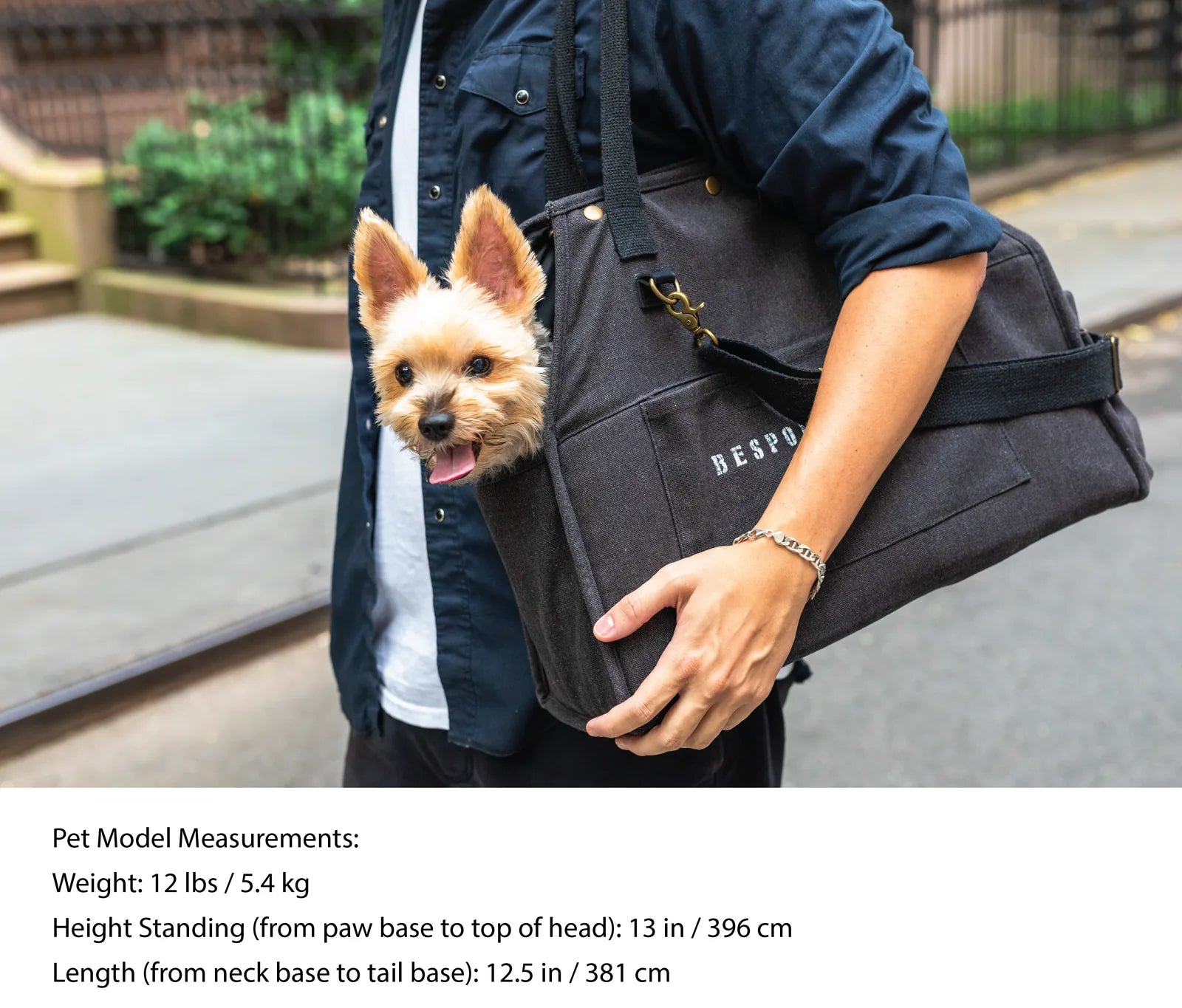 A person carries a small dog in a black, packable pet carrier with a stylish design, showcasing the BESPOKE PAWS brand.