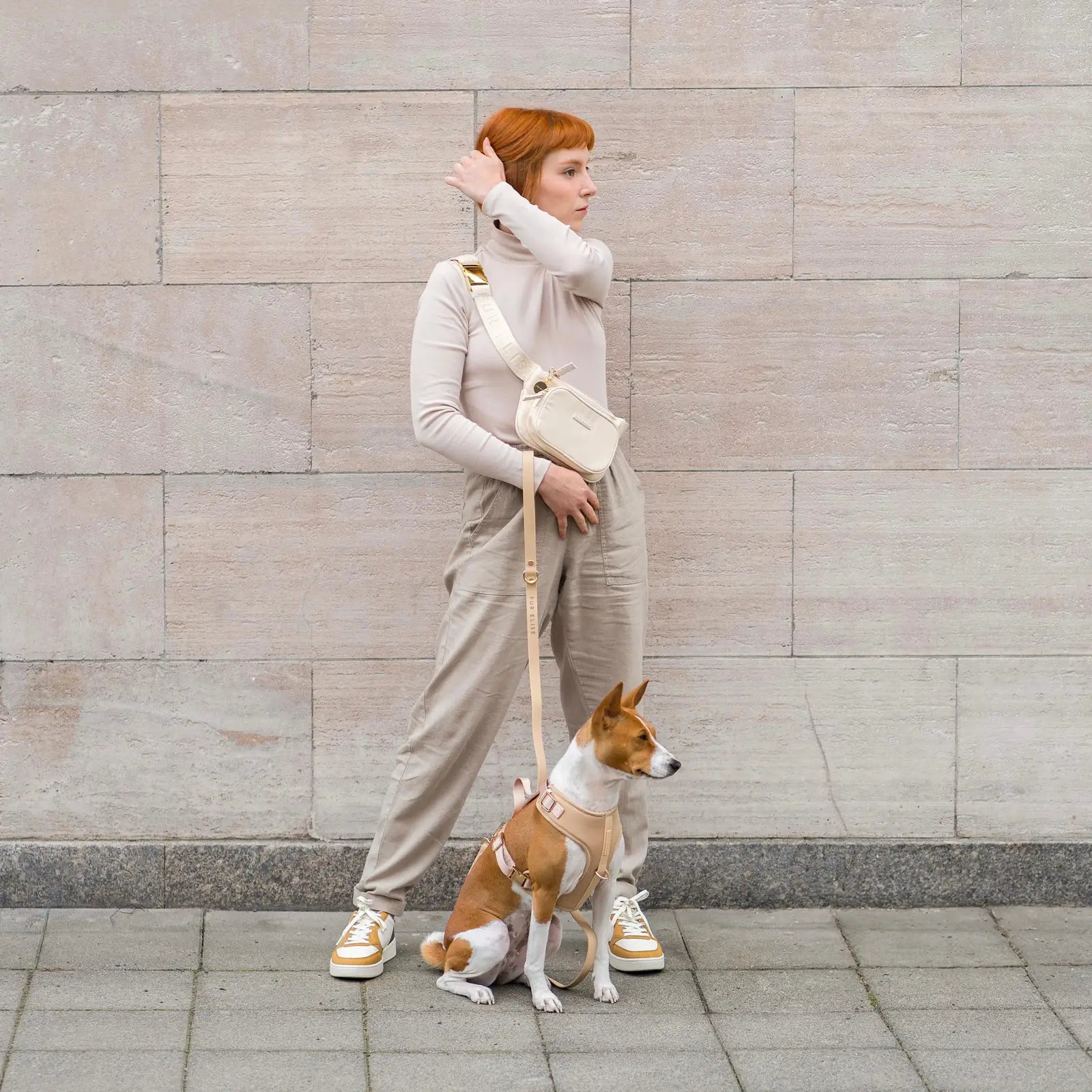 A woman in a beige outfit holds a soft beige leash attached to a dog wearing a matching harness, set against a stone wall.