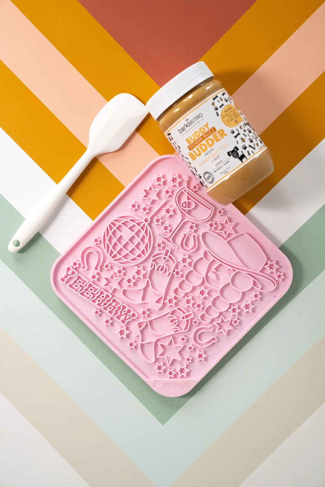 Pink silicone lick mat with star and cowboy hat designs, accompanied by a white spatula and peanut butter jar on a colorfu...
