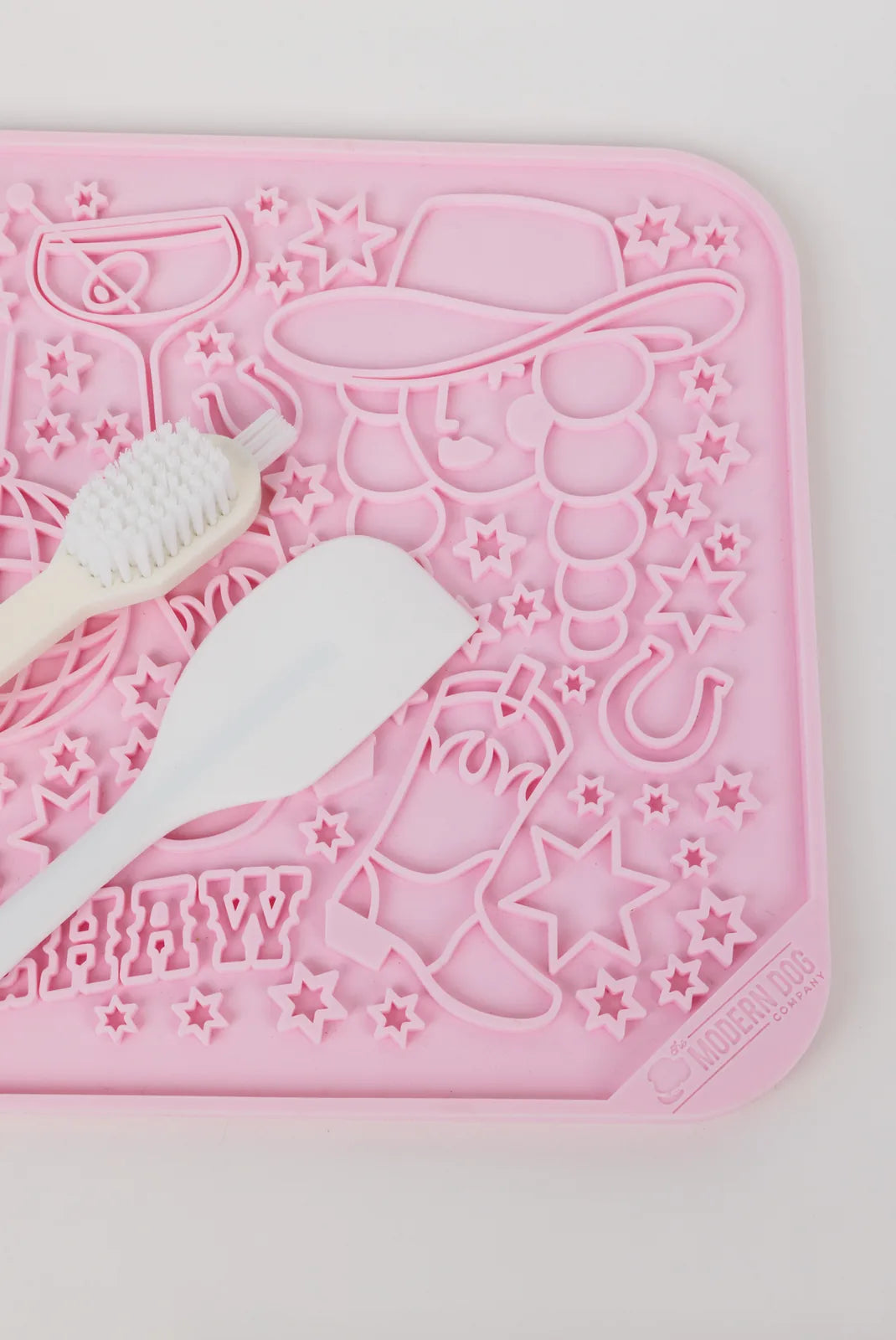 Pink silicone lick mat featuring cowgirl designs, a white spatula, and a brush for easy application and cleaning.