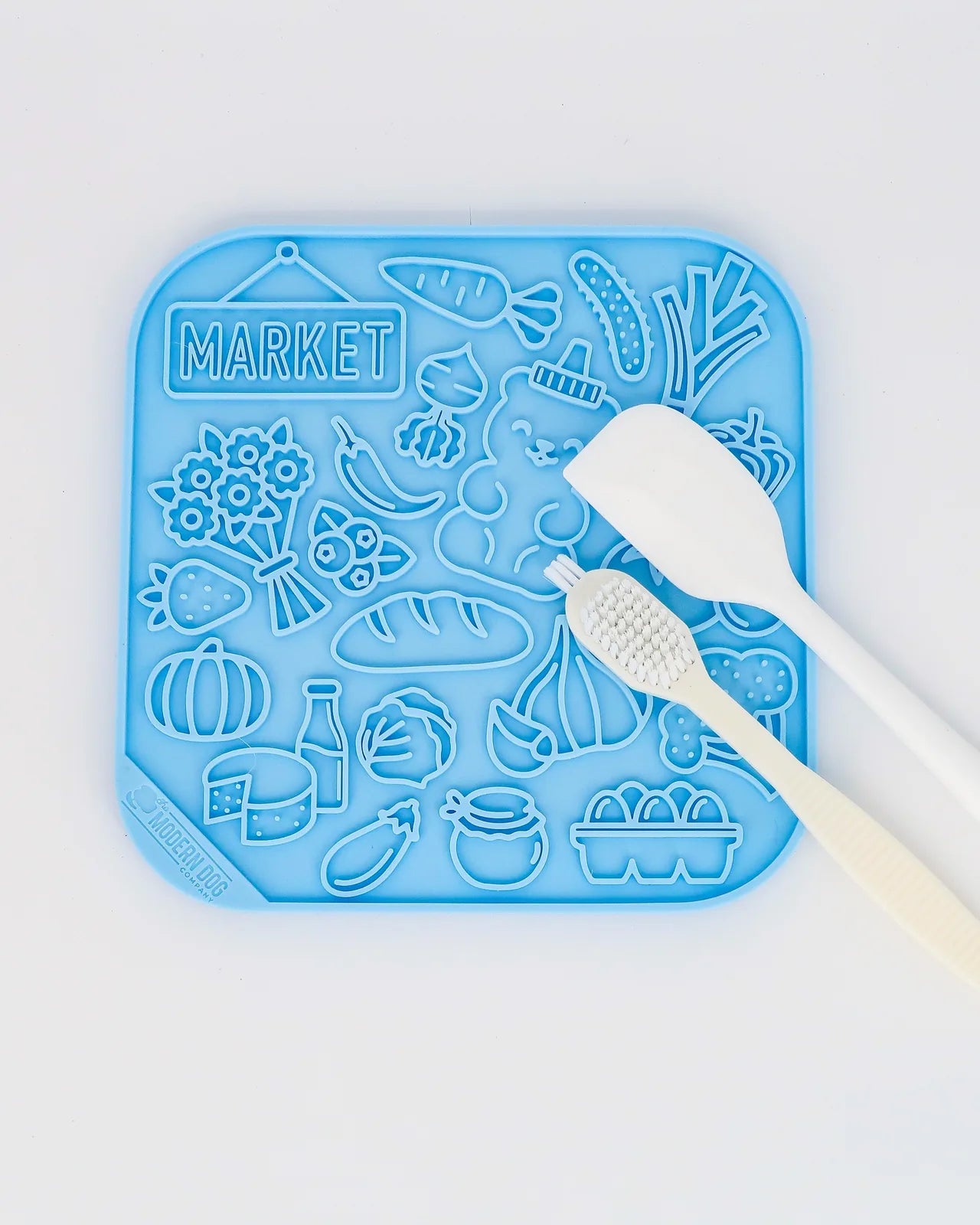 Colorful blue silicone lick mat with various food shapes, includes a spatula and scrub brush for easy use and cleaning.