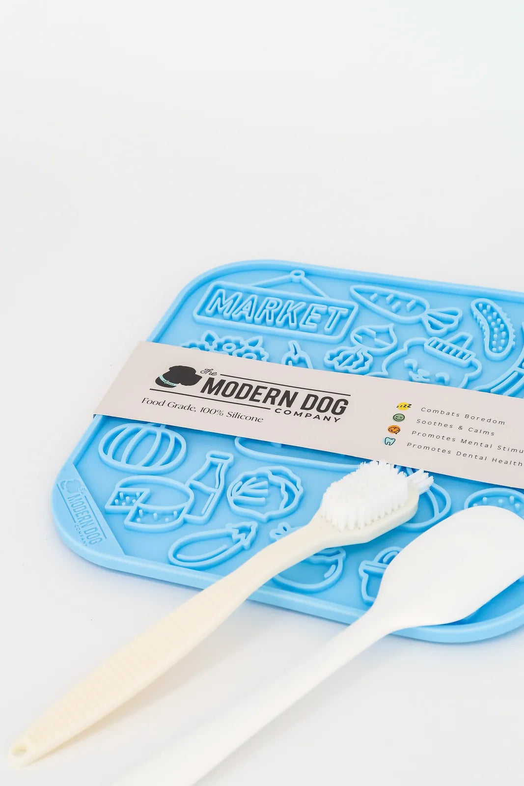 Colorful blue silicone lick mat with farmer's market designs, spatula, and scrub brush for dog feeding enrichment.