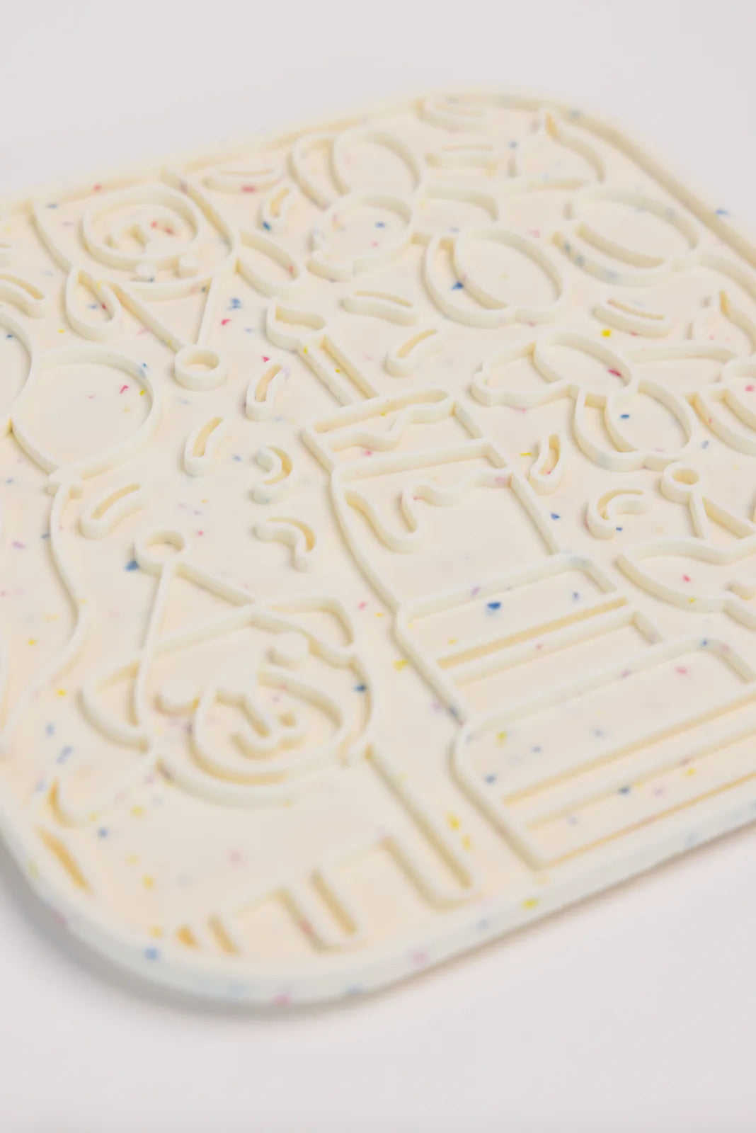 It’s a birthday pawty lick mat in cream with colorful speckles, featuring playful designs of balloons and cake.