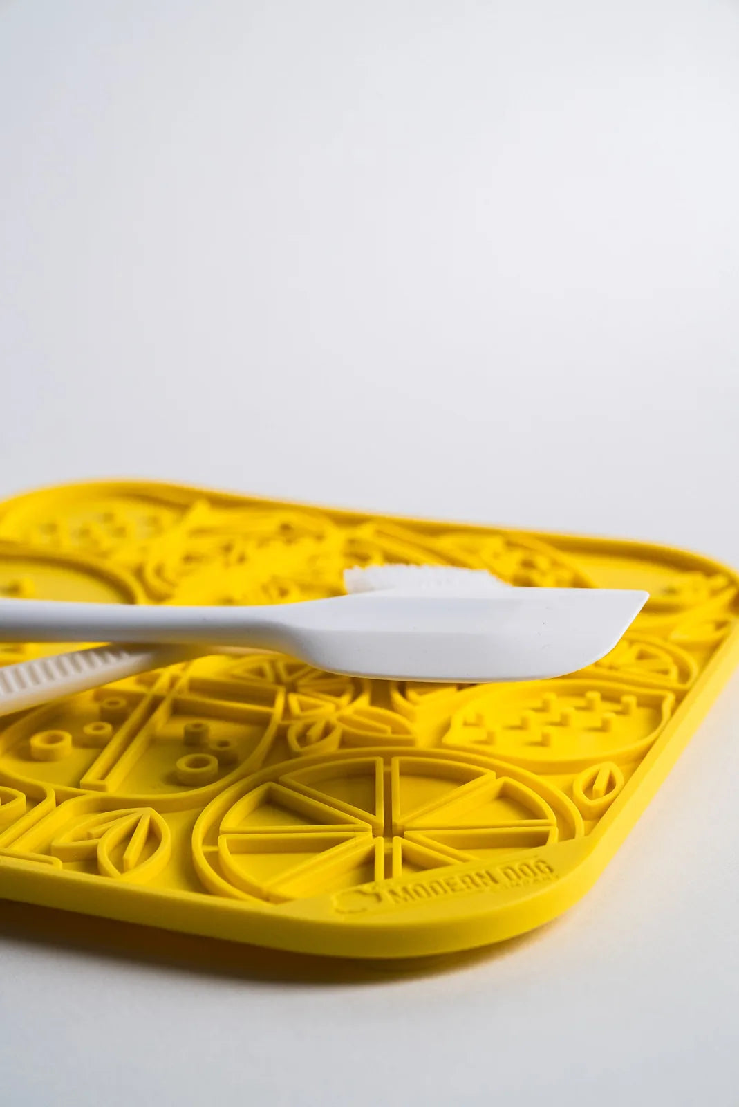 White spatula resting on a vibrant yellow food-grade silicone lick mat with a citrus pattern.
