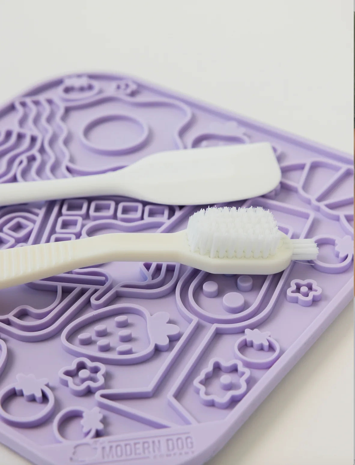 Purple silicone lick mat with textured design featuring a spatula and cleaning brush on top.