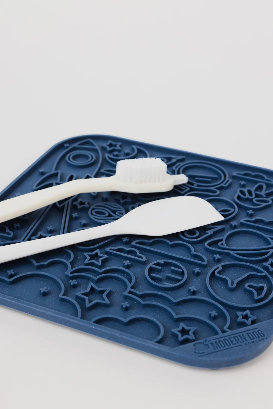 Navy blue silicone lick mat featuring a galaxy pattern, with a white spatula and brush on top for spreading treats.