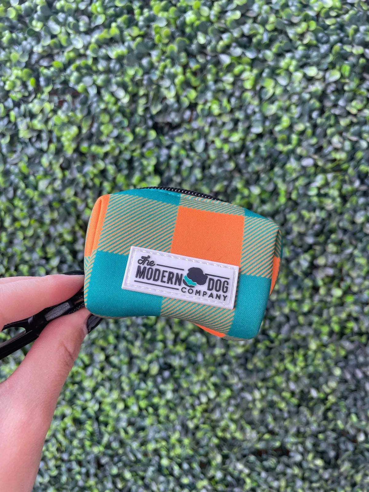 Plaid camping poop bag holder in teal and orange, featuring The Modern Dog Company logo, against a greenery backdrop.