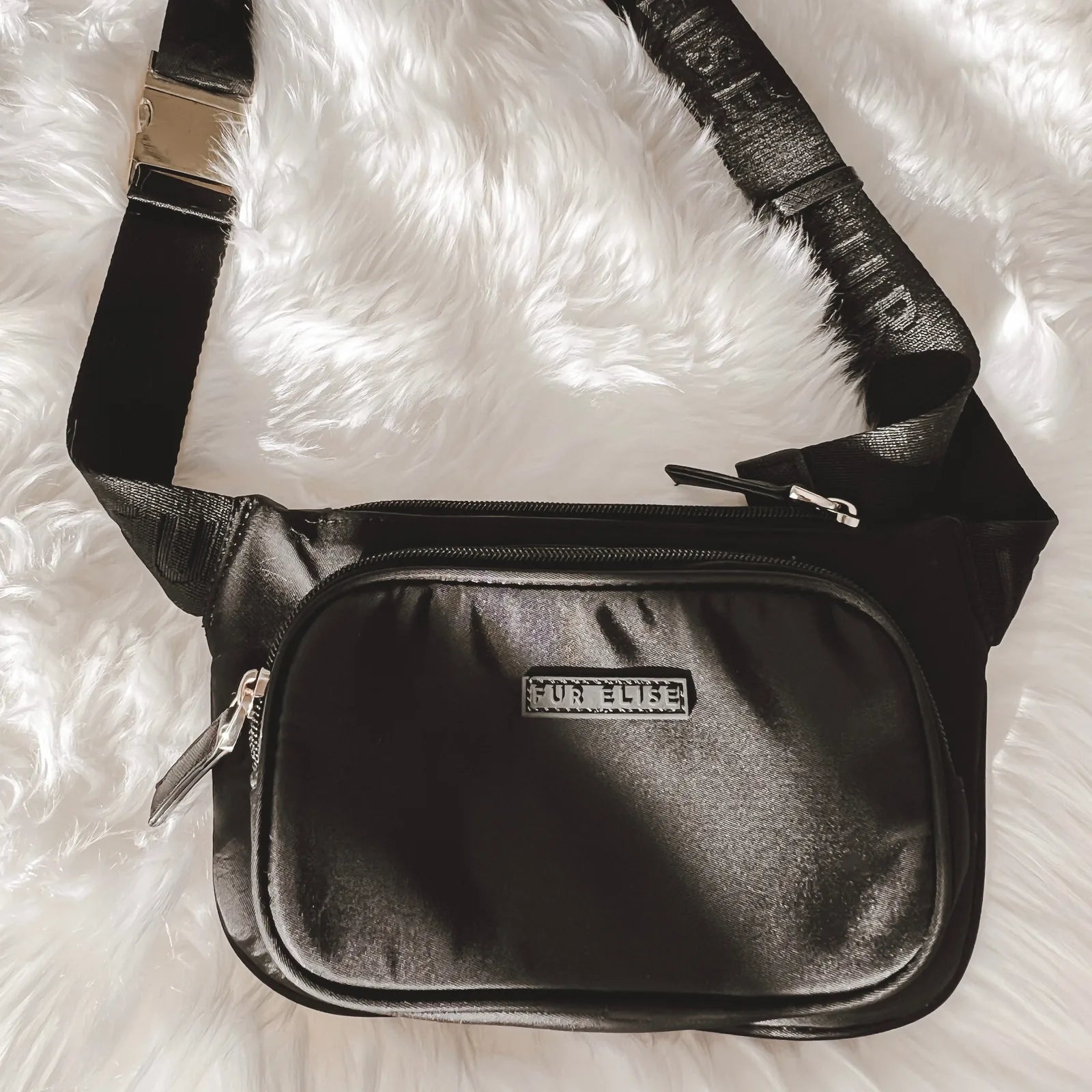 Black belt bag with shiny finish and logo, featuring a zippered compartment and adjustable strap on plush white fur.