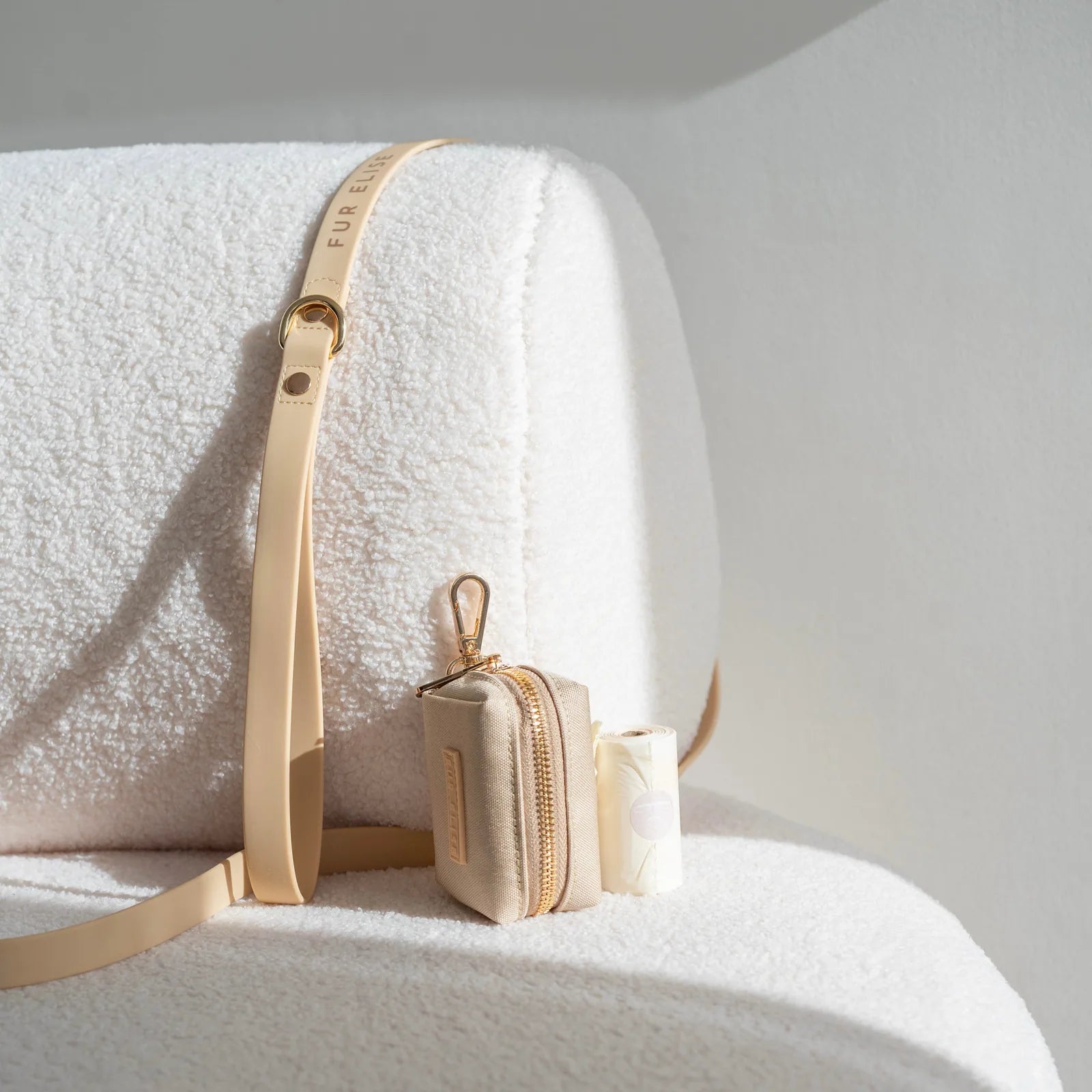 Beige leash with gold hardware alongside a compact beige pouch and biodegradable dog waste bags, on a textured white surface.