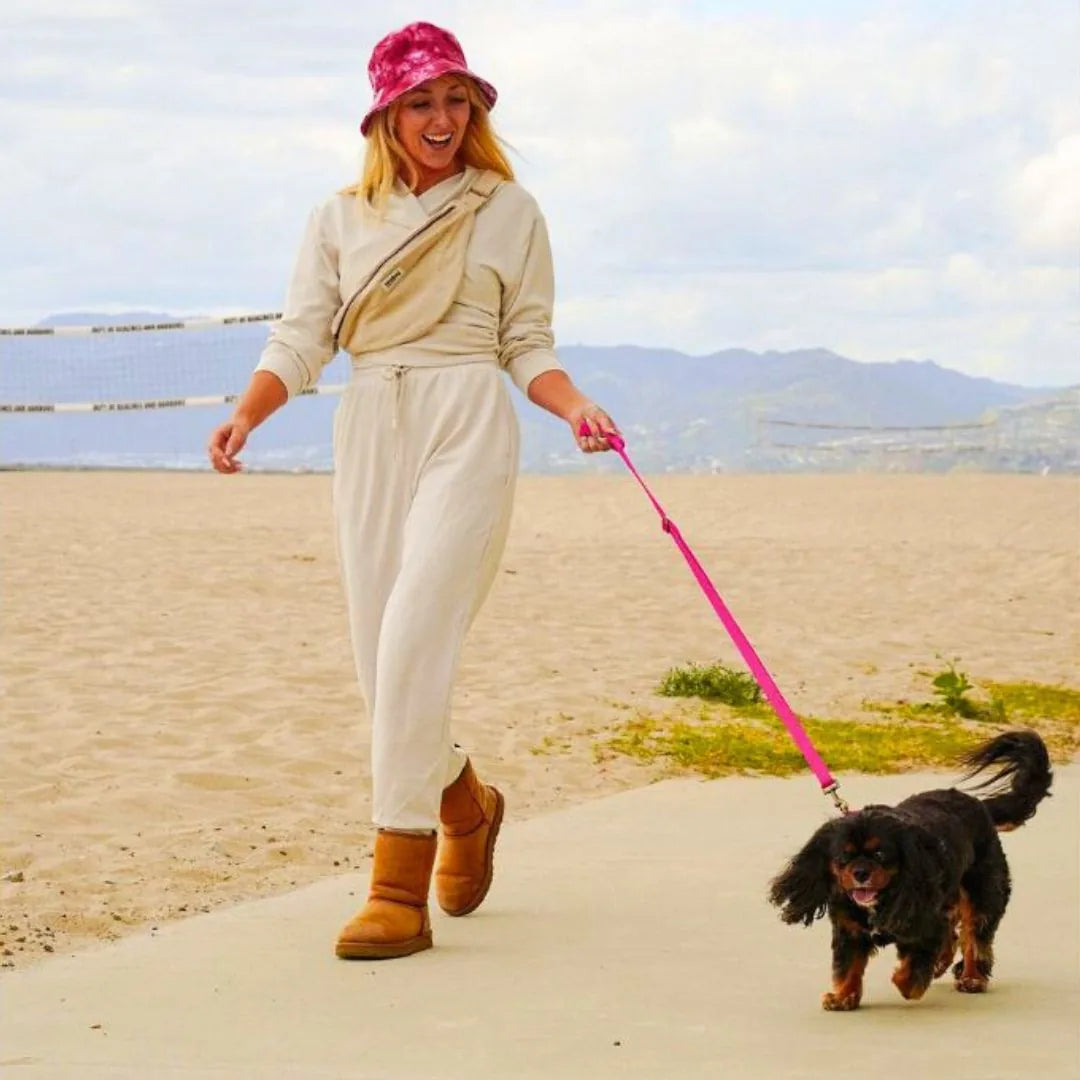 Woman in a light jumpsuit walks a small black dog on a pink hemp cotton leash along a sandy beach path.
