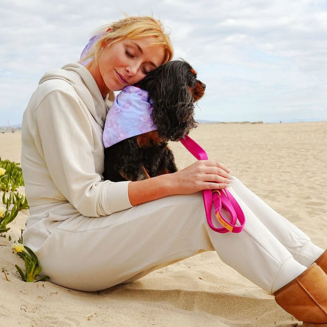 Woman cuddles a black dog on the beach, showcasing a vibrant pink hemp cotton leash and a pastel tie-dyed scarf.