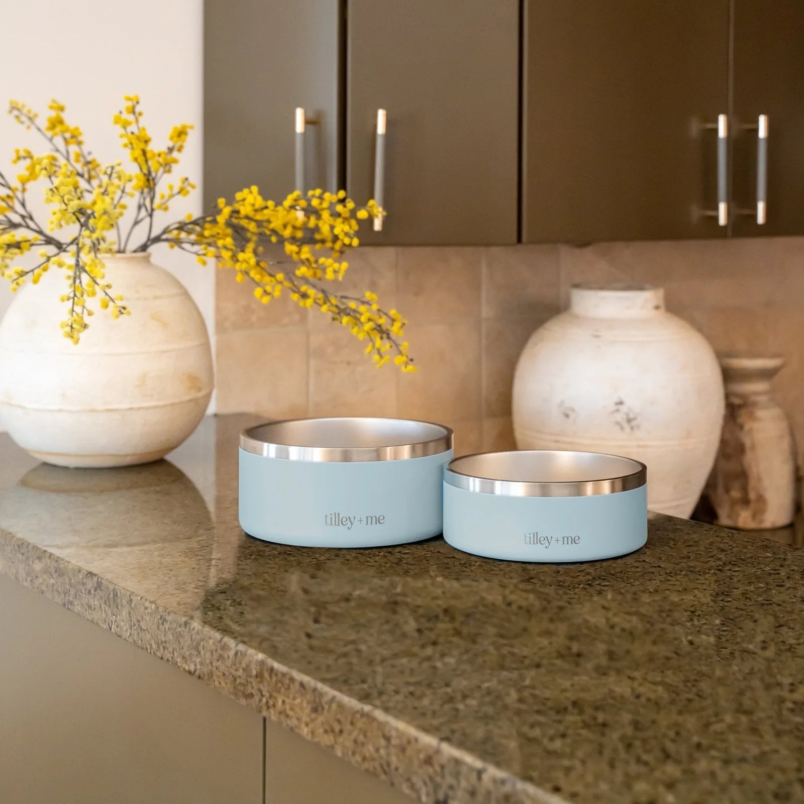 Light blue stainless steel pet bowls with non-slip bases sit on a granite countertop, featuring the "tilley + me" logo.