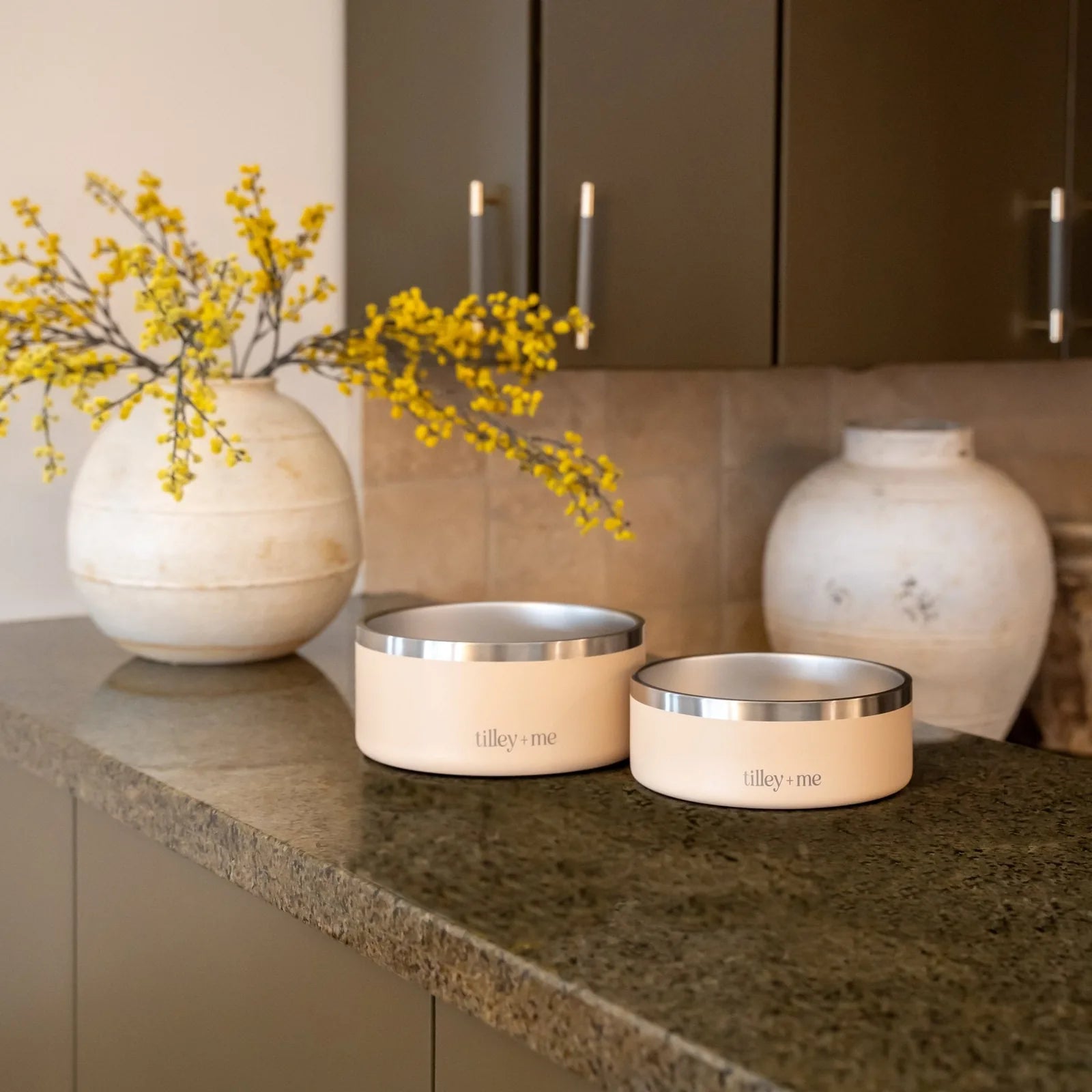 Terracotta Rose stainless steel pet bowls with non-slip bases, displayed on a countertop with yellow flowers in a vase.