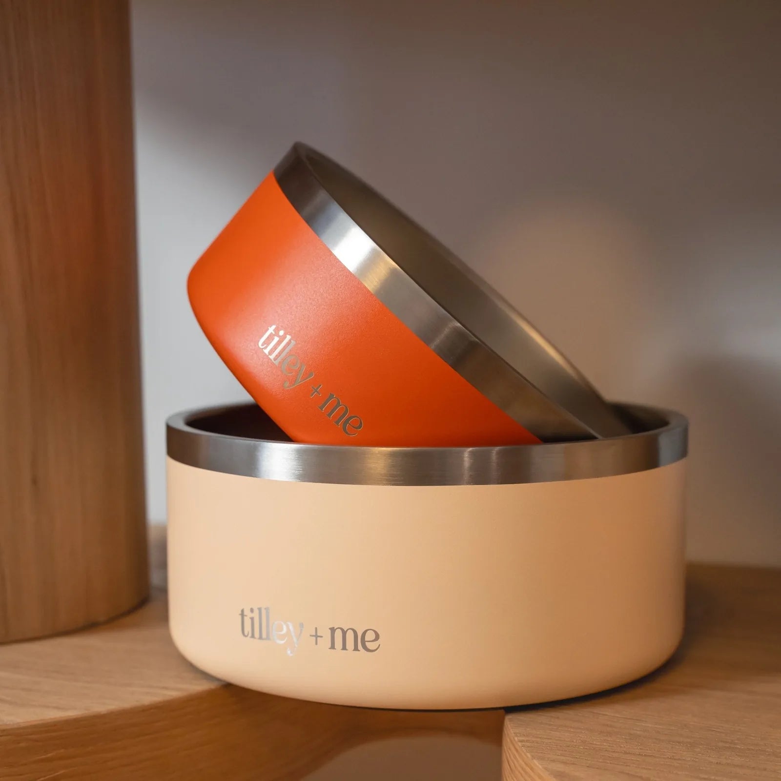 Non-slip stainless steel pet bowls stacked, featuring a terracotta orange bowl on top of a soft beige bowl with "tilley +...