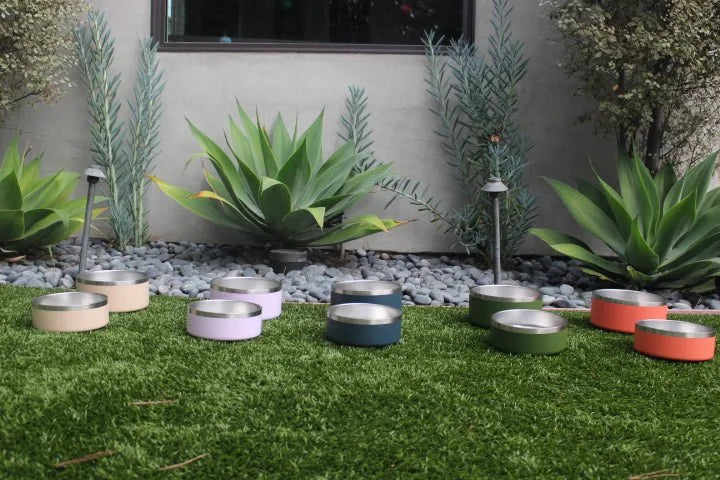 Stainless steel dog bowls in various colors, including terracotta, lilac, and green, arranged on grass with greenery in th...