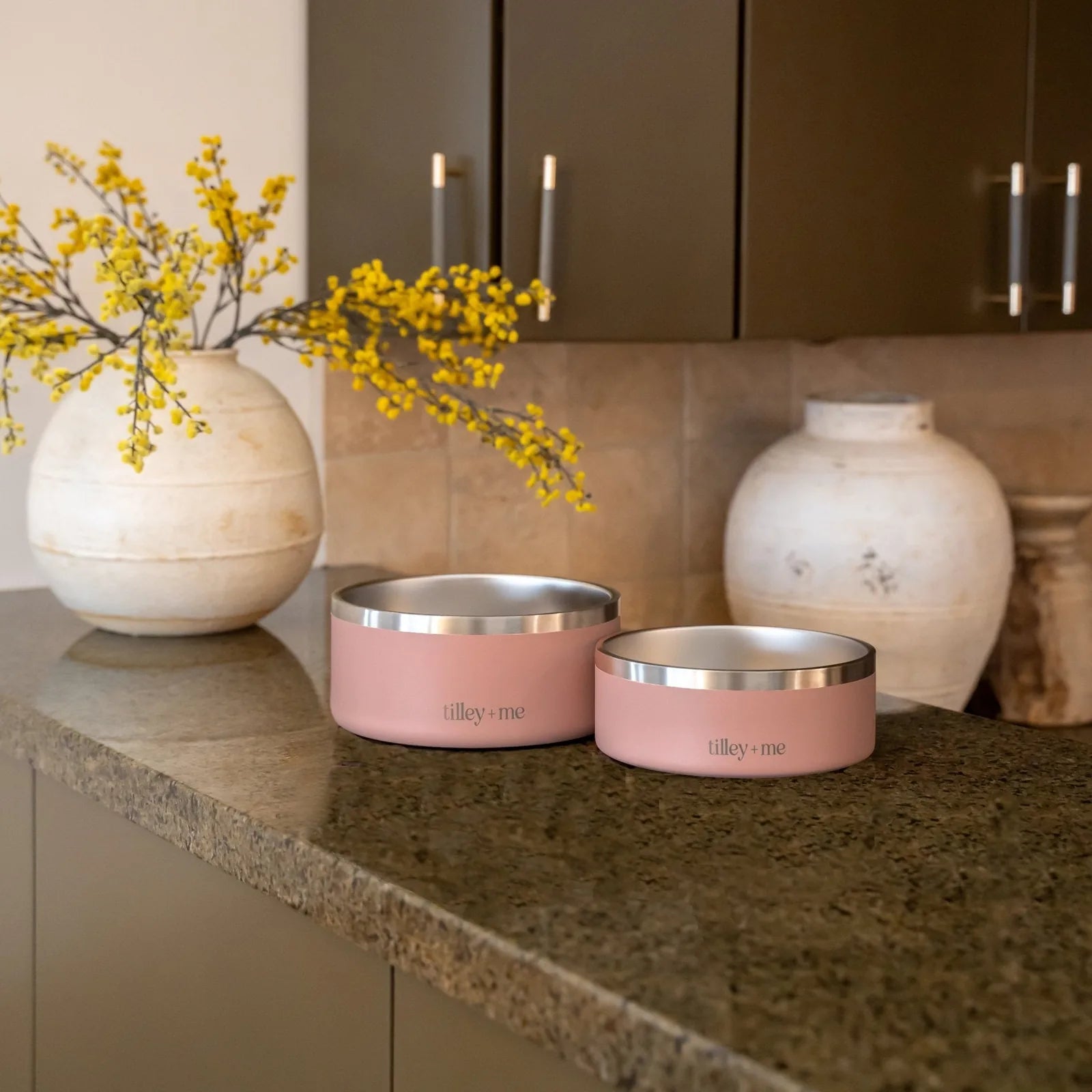 Terracotta Rose stainless steel pet bowls with non-slip bases on a granite countertop, surrounded by decorative vases.