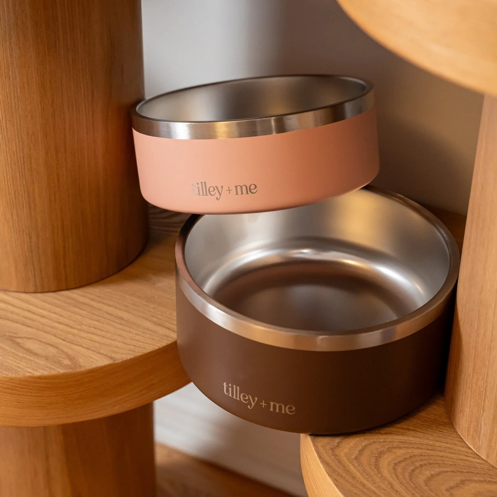 Stainless steel pet bowls in Terracotta Rose and Burnt Umber Brown with non-slip bases on a wooden shelf.
