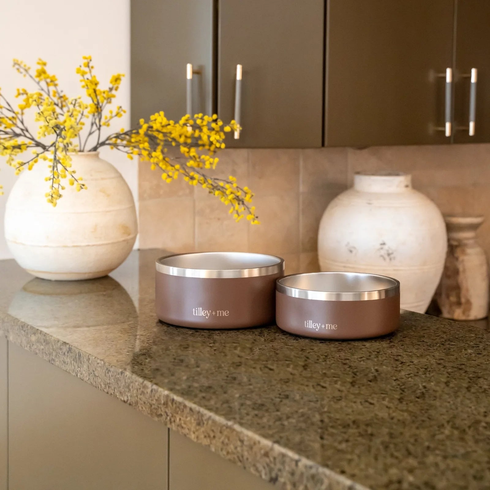 Two Tilley + Me stainless steel pet bowls in Burnt Umber Brown, one large and one small, on a granite countertop with deco...