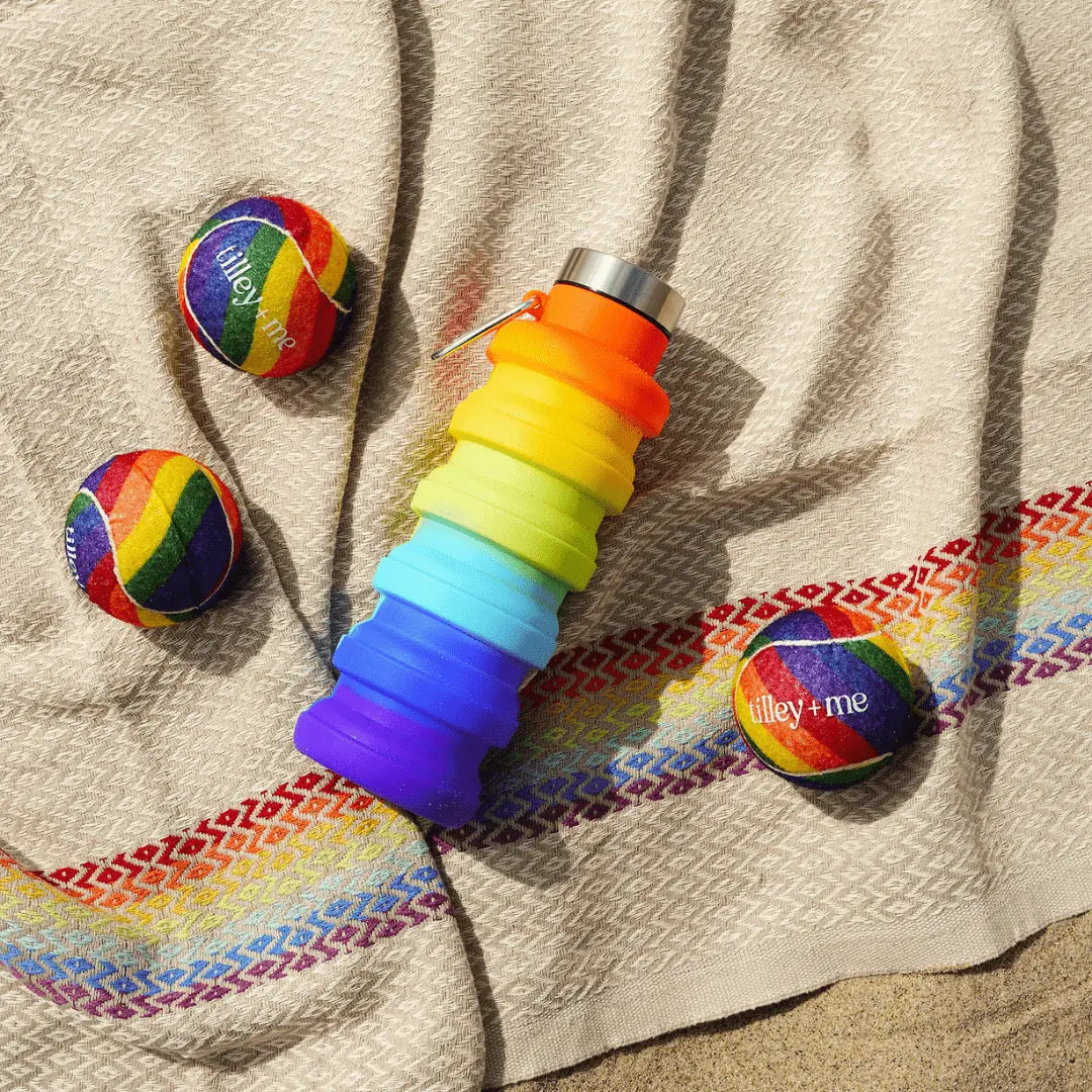 Colorful Tilley + Me tennis balls in rainbow patterns next to a vibrant collapsible water bottle on a textured blanket.