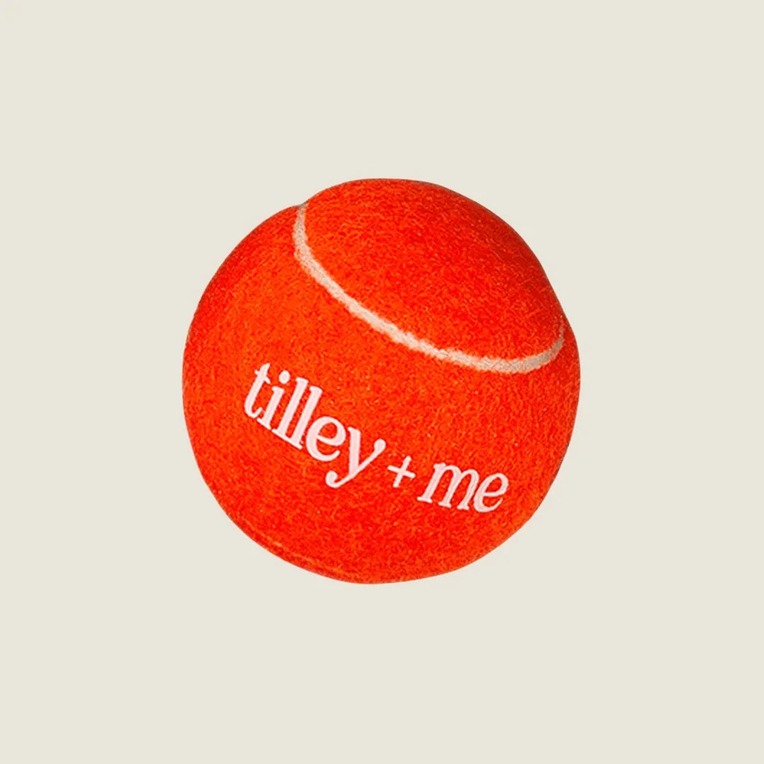 Bright orange tennis ball for dogs with white stitching and "tilley + me" logo, made from sustainable materials.