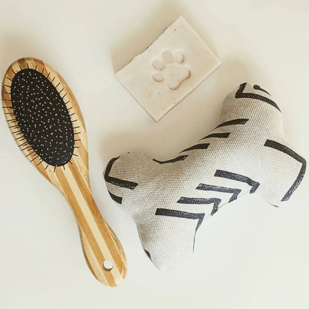 Two-sided dog brush with bamboo handle, paired with a bone-shaped toy and paw-print soap on a light background.