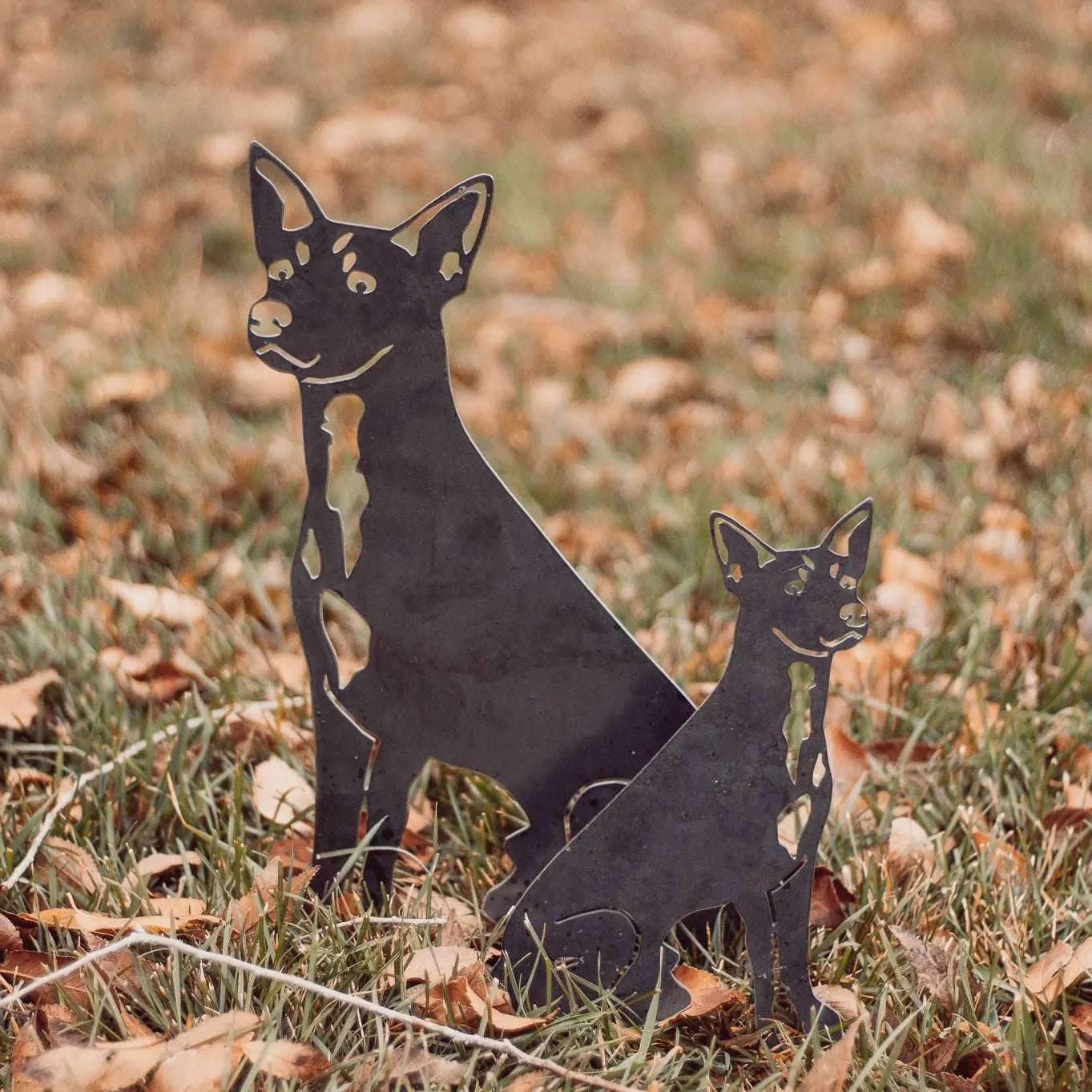 Toy Fox Terrier metal silhouettes in Corten steel, set against a backdrop of brown grass and fallen leaves.