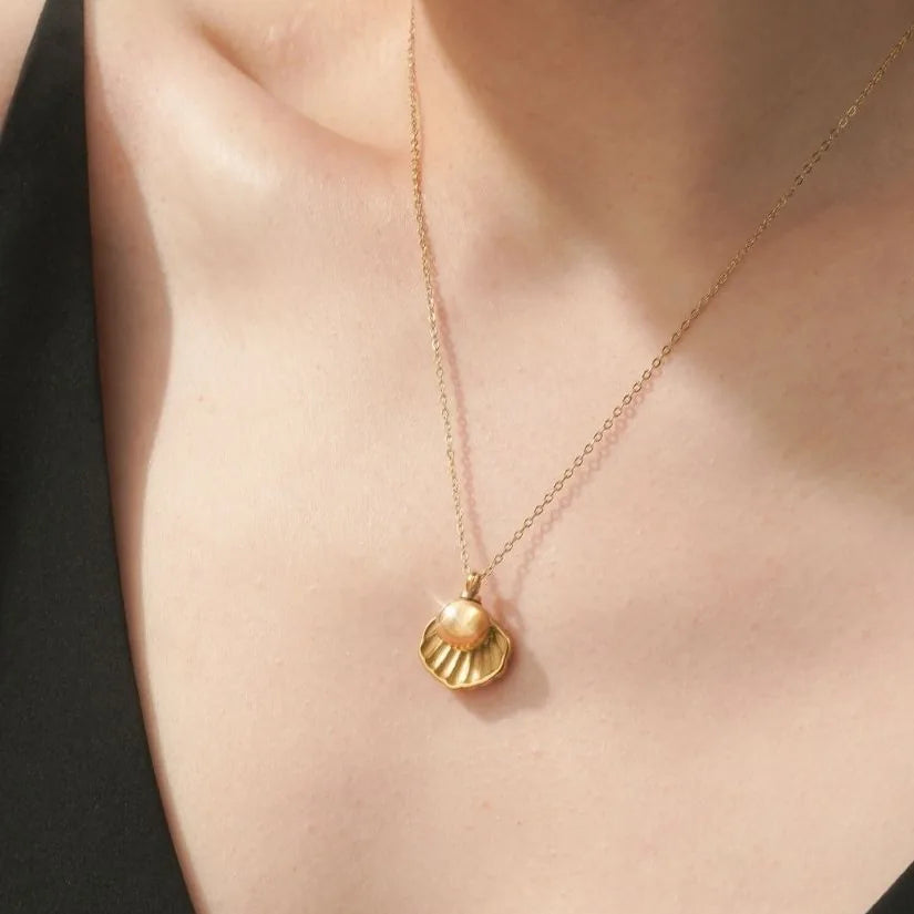Gold memorial necklace featuring a shell design, delicately holding a pearl, worn on a chain against bare skin.