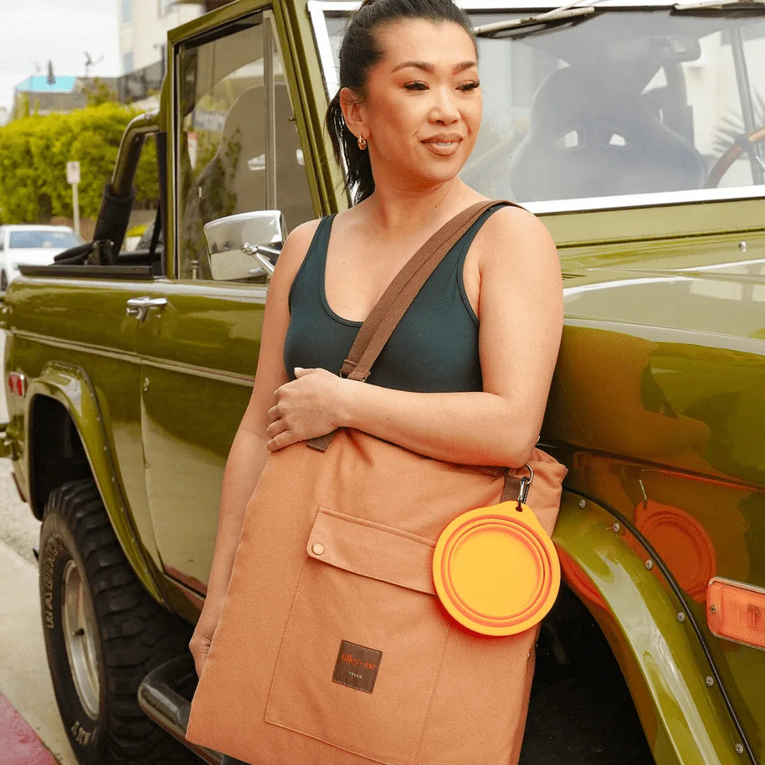 Woman holding a brown canvas travel mat with a pocket, featuring an orange dog dish, beside a vintage green jeep.