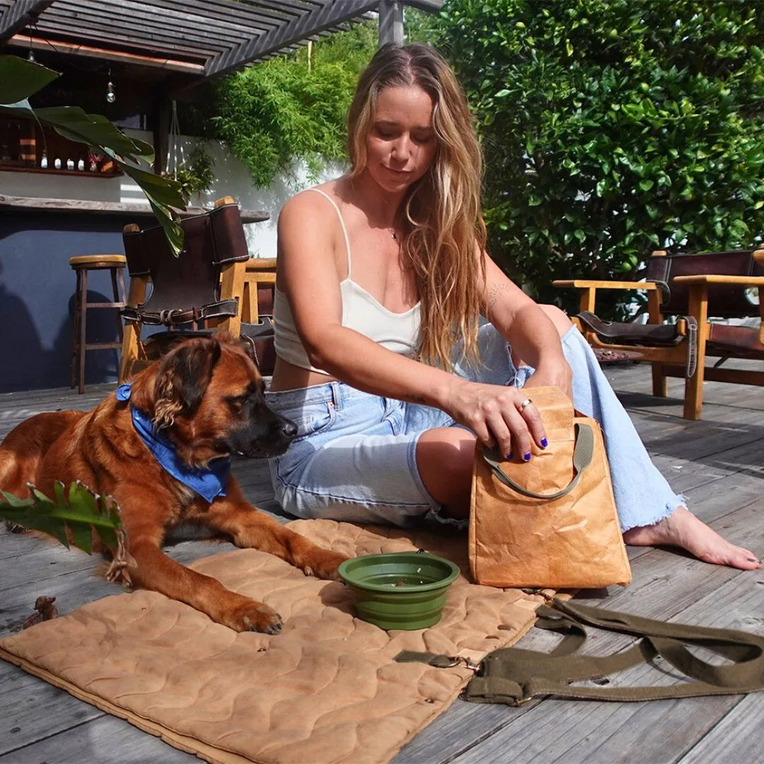 A relaxed dog rests on a tan, quilted travel mat beside a woman sitting on a wooden deck with outdoor greenery.