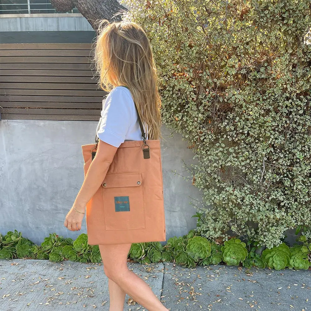 Lightweight portable canvas mat in terracotta color, worn as a messenger bag, set against a leafy backdrop.