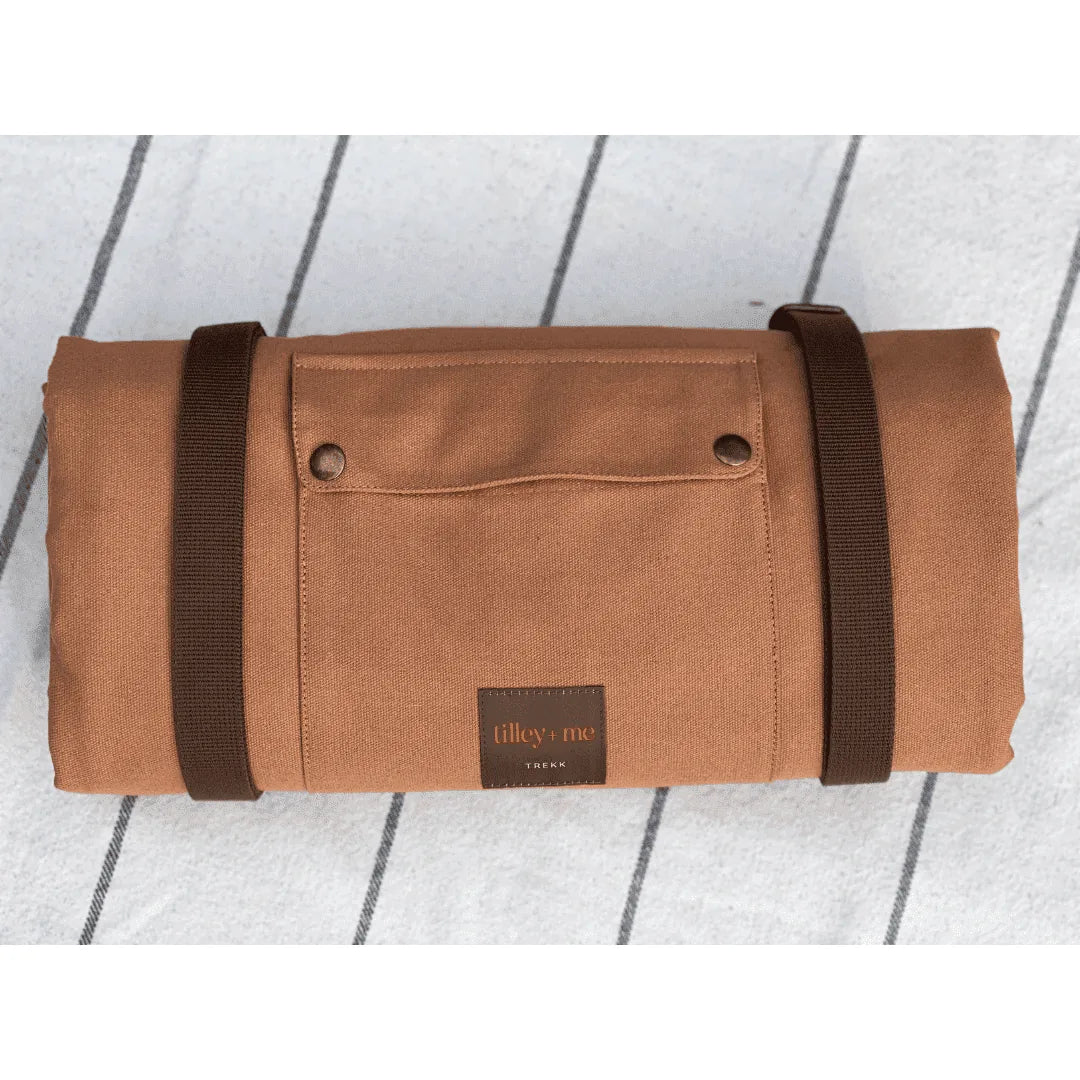 Brown canvas Trekk Convertible Travel Mat with dark straps and a logo, rolled up on a light striped surface.