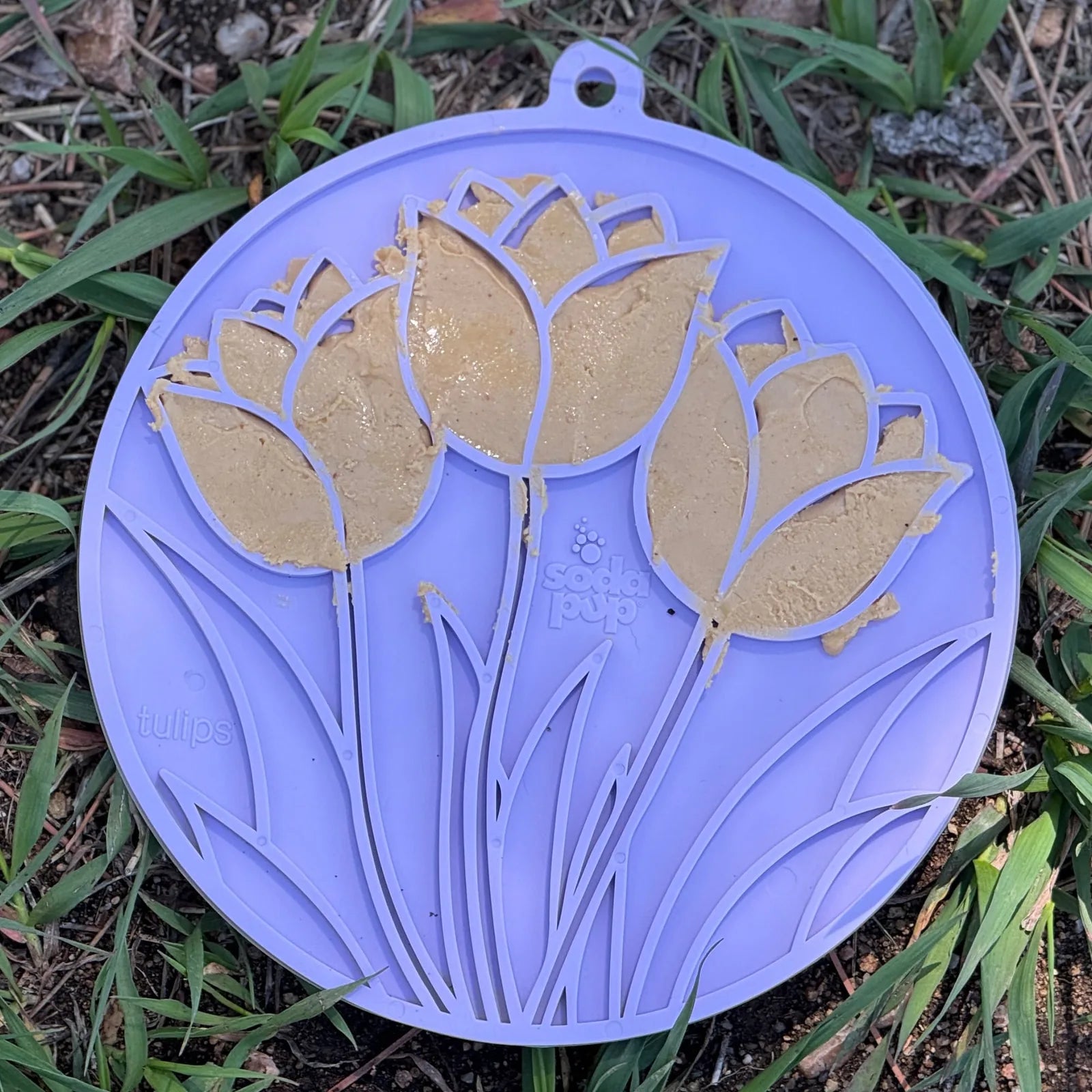 Pastel purple tulip design lick mat with treat spread, set on grass, showcasing textured surface for dogs and cats.