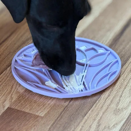 Black dog licking a pastel purple Tulip Design eMat on a wooden floor, featuring textured floral patterns for treat enrich...