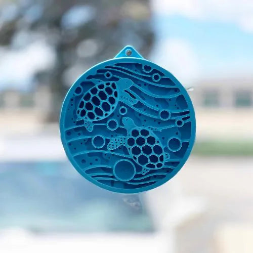 Turtle-shaped blue lick mat with suction cups, featuring varied textures for small pets, set against a blurred outdoor bac...