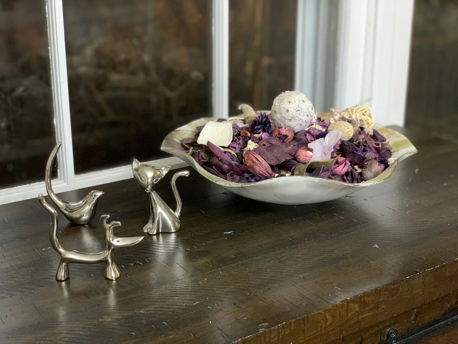 Modern silver figurines of a dog, cat, and bird, displayed among decorative potpourri in a bowl, on a wooden surface.