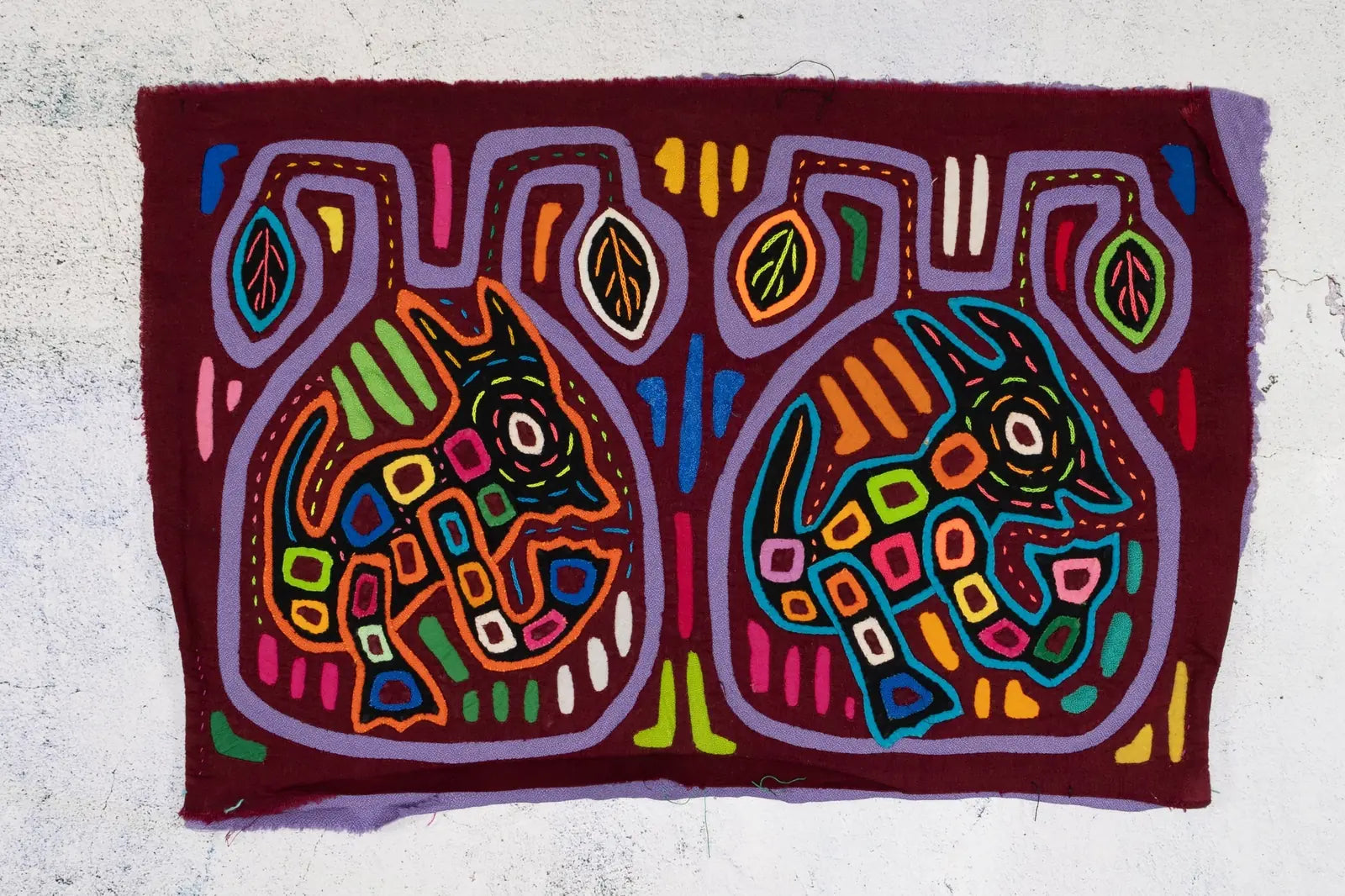 Colorful hand-stitched textile art featuring two playful puppy designs in vibrant hues against a deep maroon background.