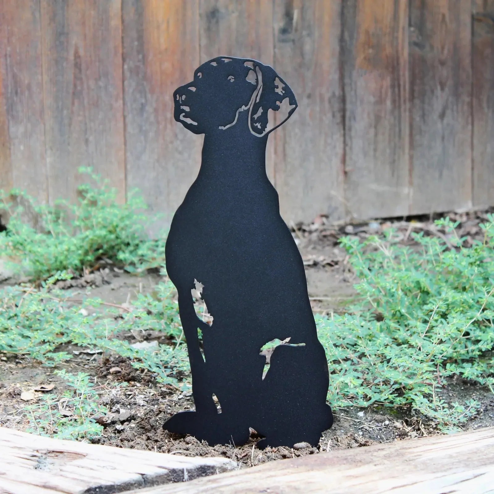 Black metal silhouette of a Vizsla dog sitting, surrounded by greenery and rustic wood background.