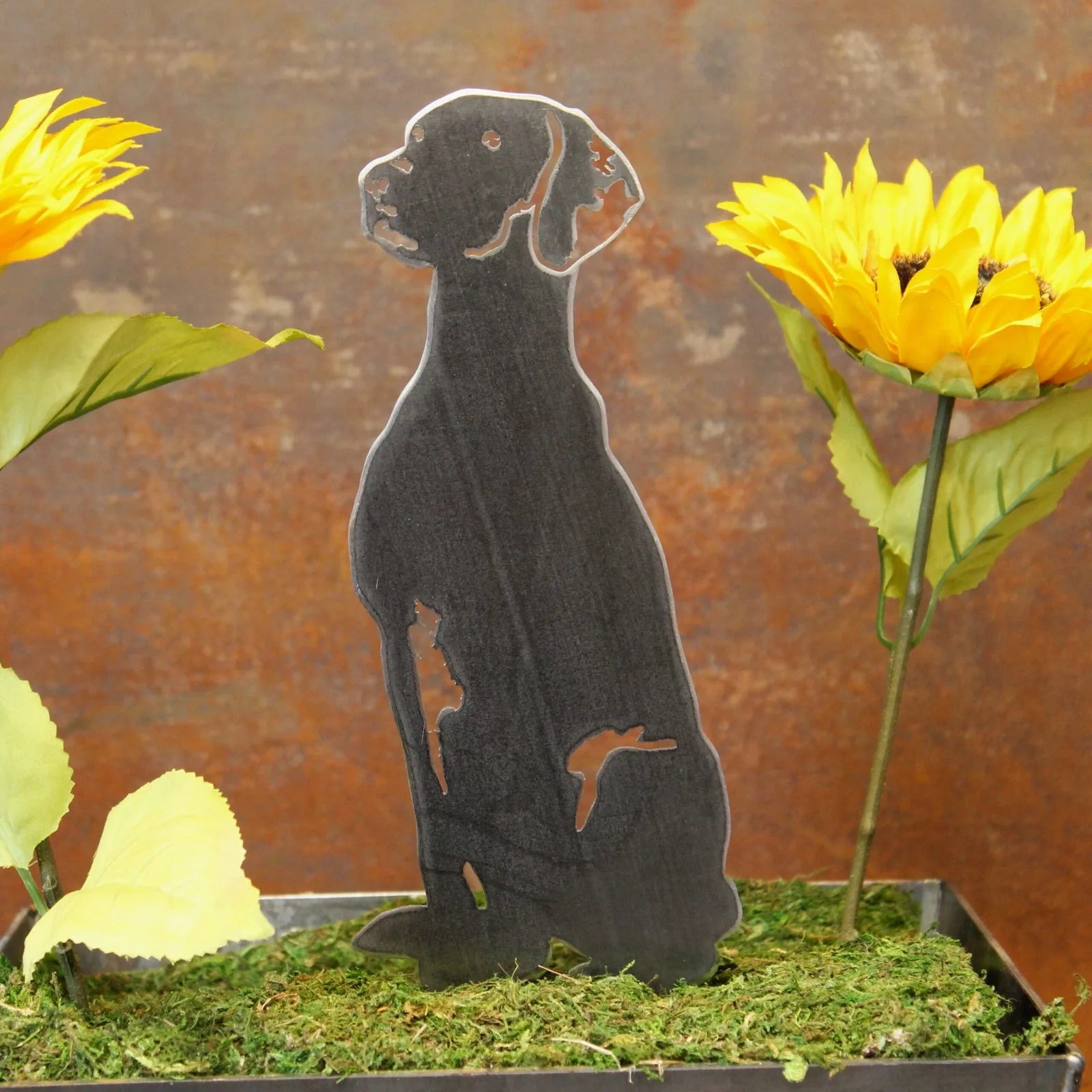 Black Vizsla metal silhouette standing among yellow flowers and green moss, showcasing sleek design and sturdy material.