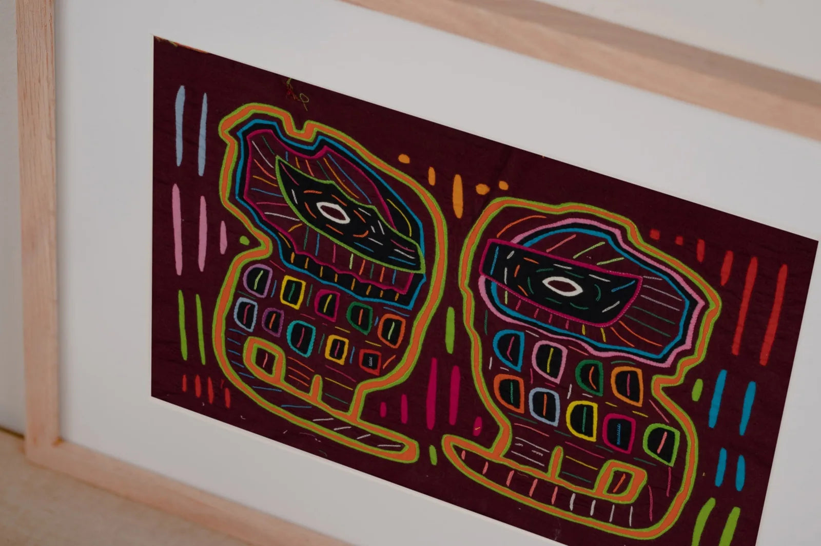 Colorful hand-stitched Mola tapestry featuring abstract dog shapes on a deep burgundy background, framed in light wood.