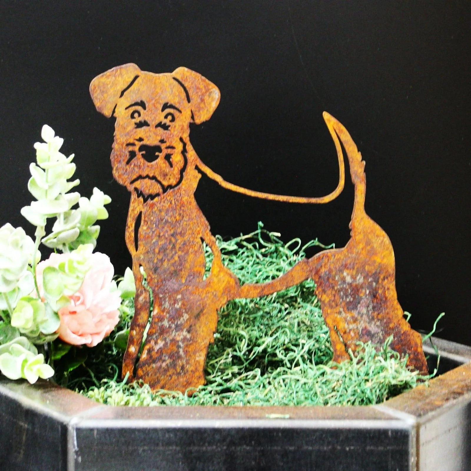 Welsh Terrier metal silhouette in rusted Corten steel, featuring detailed dog design, surrounded by green moss and flowers.