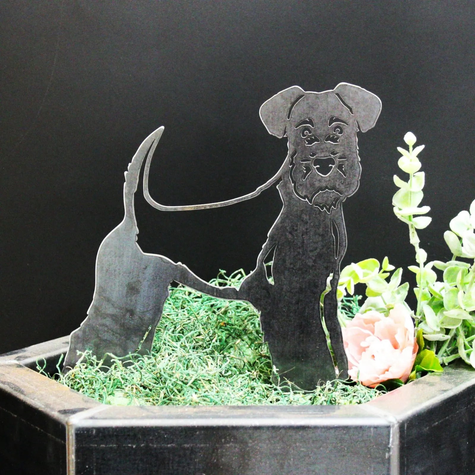 Welsh Terrier metal silhouette in black, showcasing a standing dog design, placed among greenery and flowers.