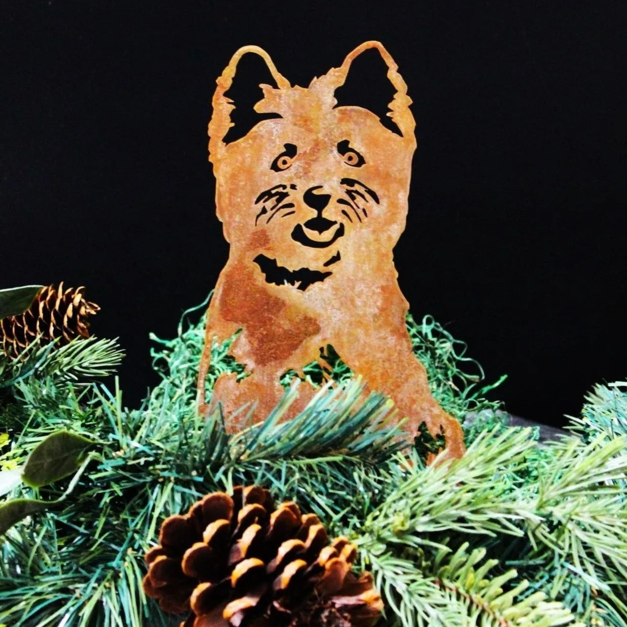 Rusty Corten steel silhouette of a West Highland White Terrier among green foliage and pinecones, ideal for outdoor decor.