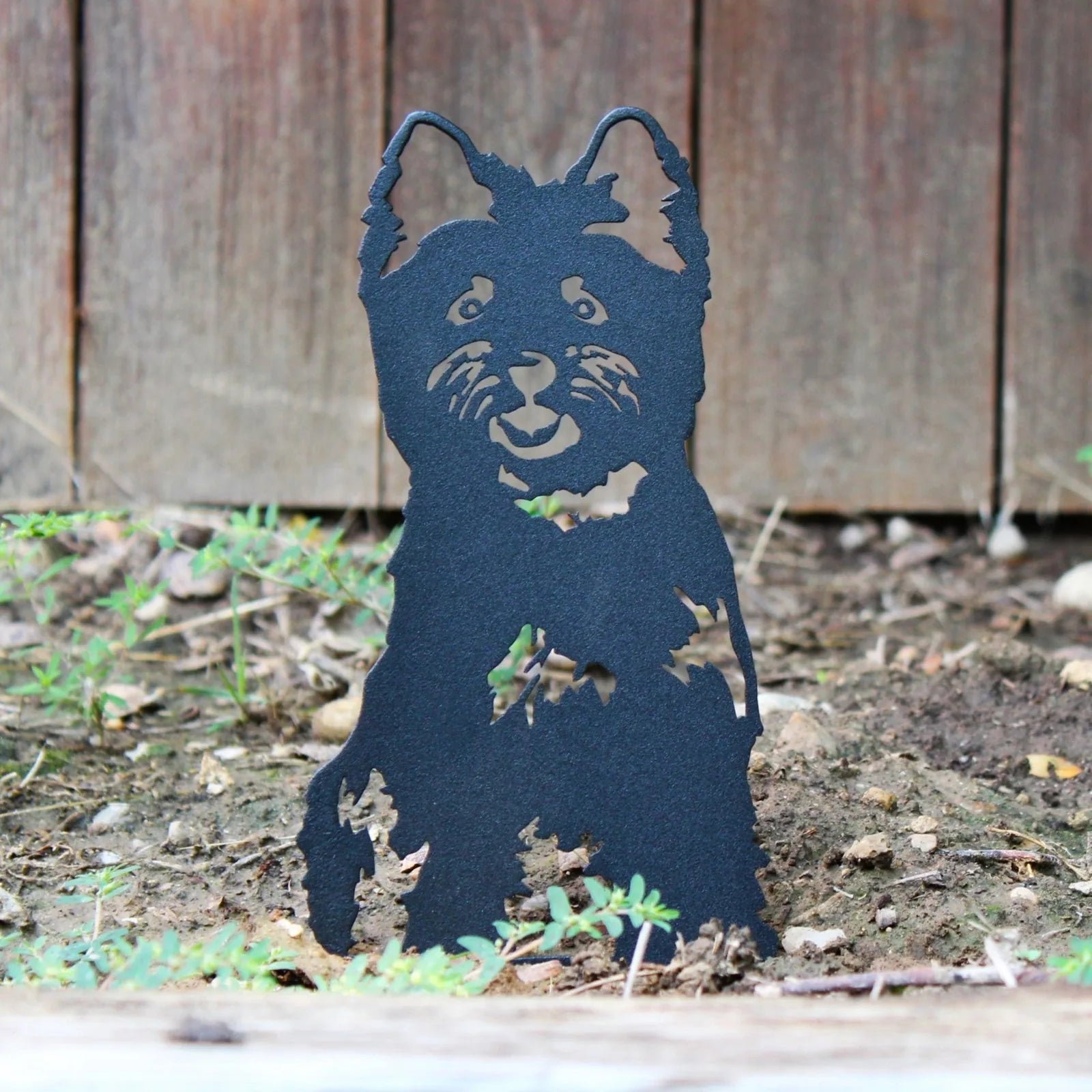 Black metal silhouette of a West Highland White Terrier, detailed with a smiling face, set against a natural garden backgr...
