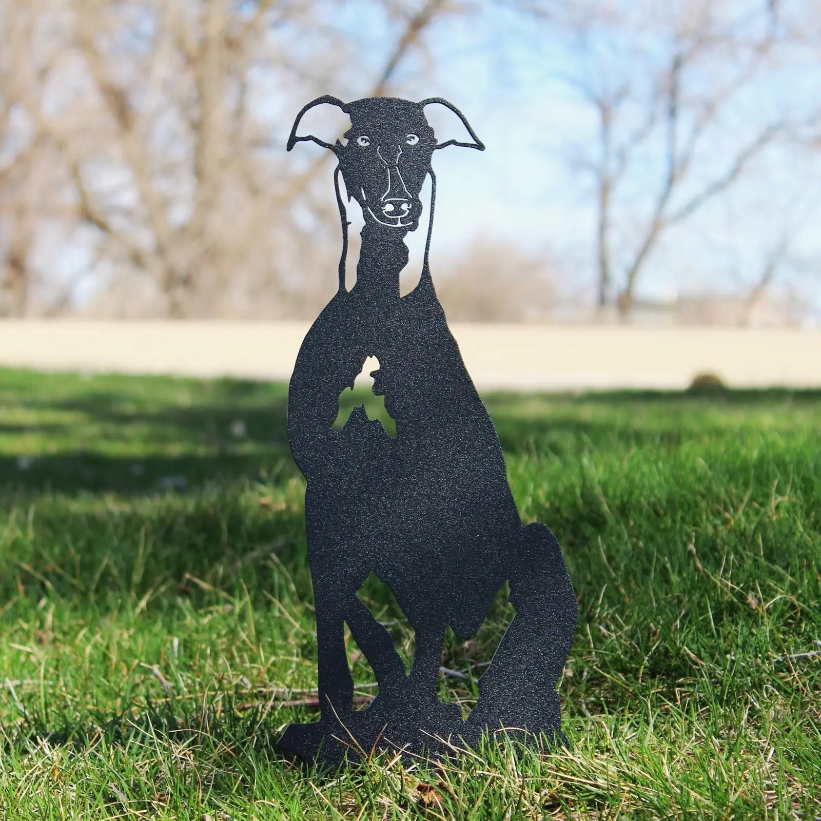Black metal silhouette of a Whippet sitting on green grass, featuring a detailed design and natural outdoor backdrop.