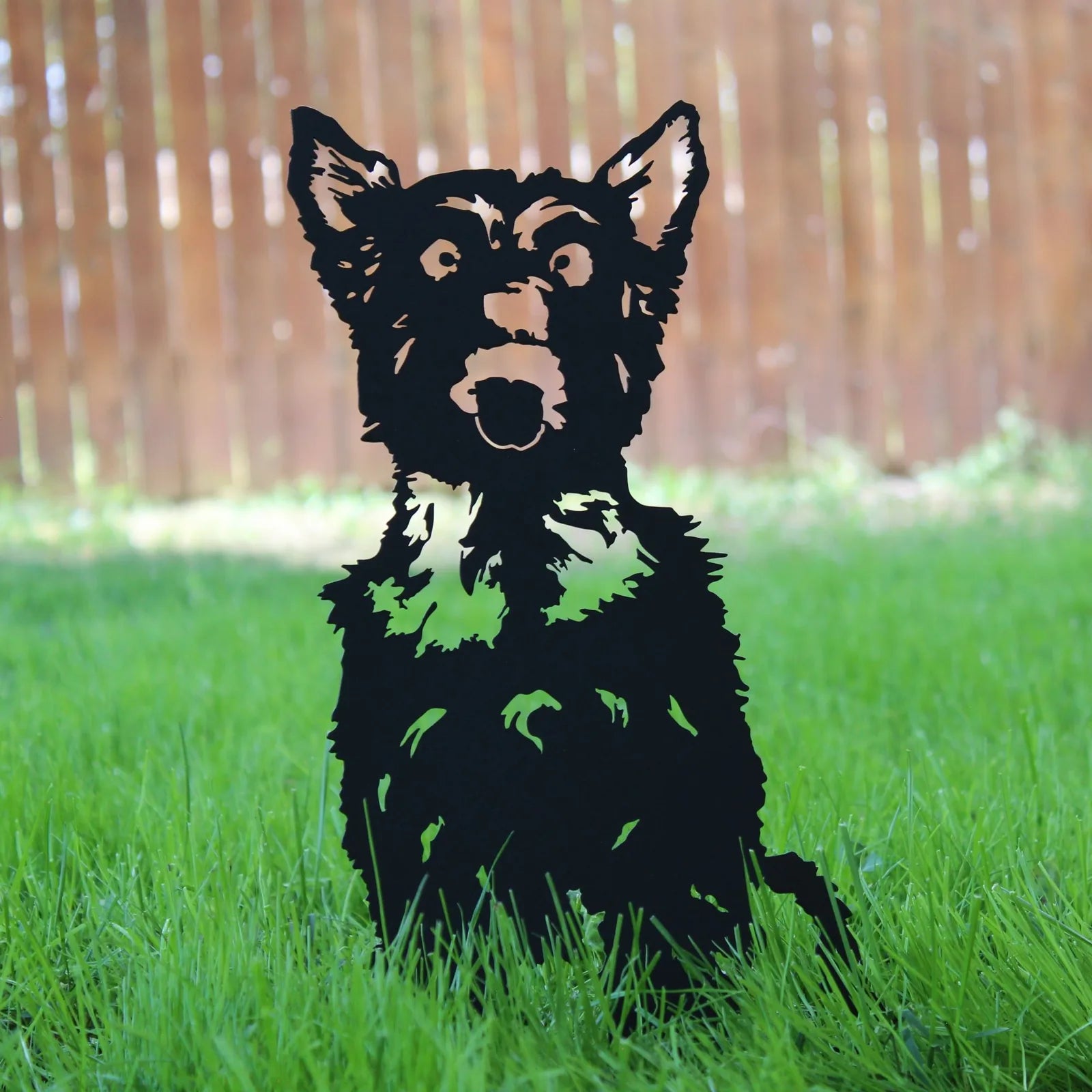 Black metal silhouette of a Yorkshire Terrier on green grass, set against a wooden fence in the background.