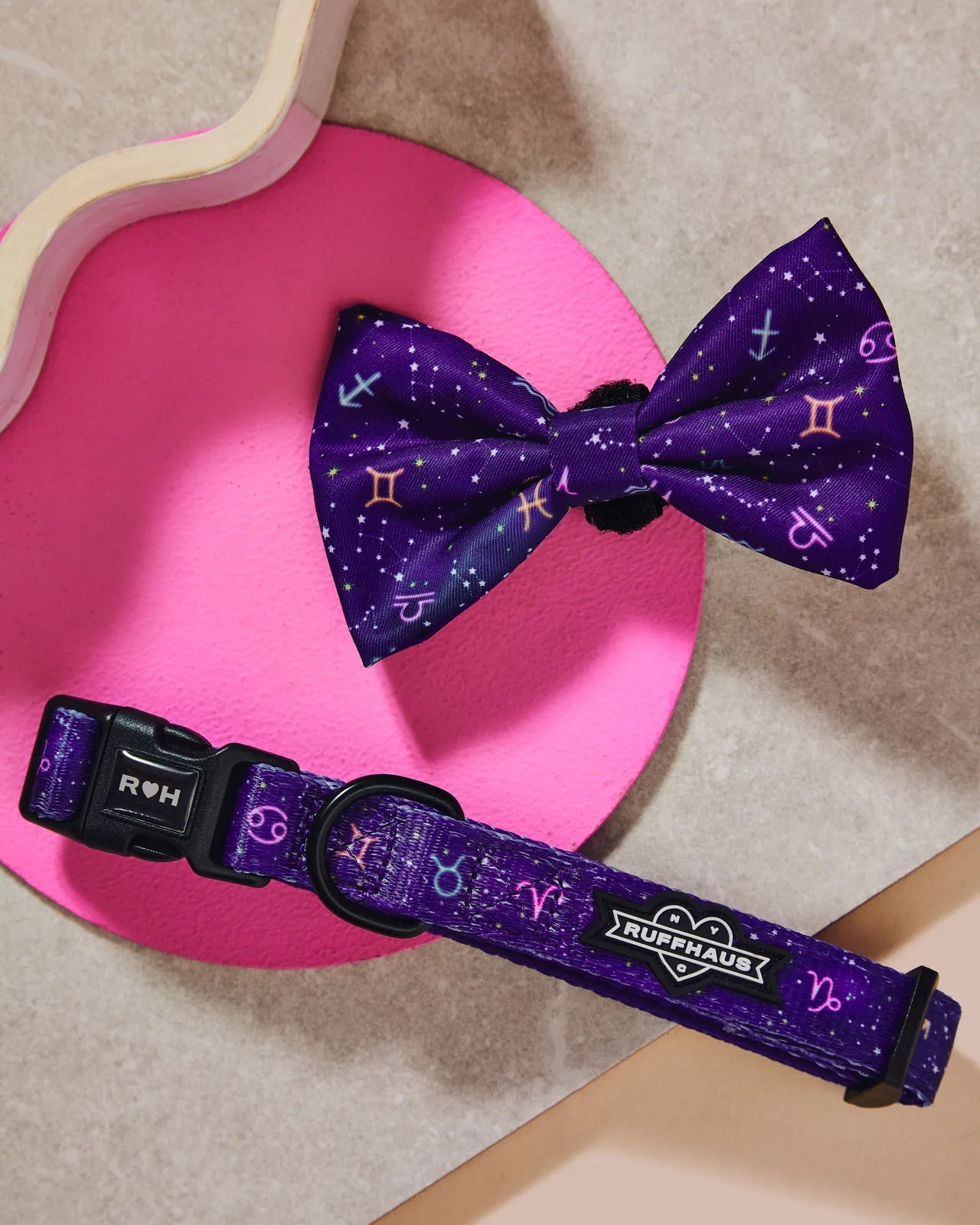 Purple dog collar with zodiac designs and a matching bowtie, set against a pink circular background.